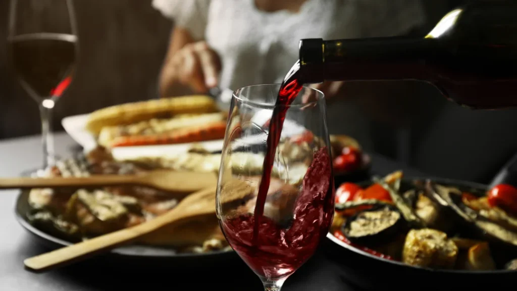 Persona sirviendo vino tinto en una copa, con un ambiente de vino y comida.