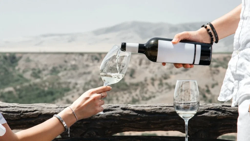 Primer plano de una persona sirviendo vino blanco desde una botella con etiqueta blanca en una copa, con un paisaje montañoso de fondo.