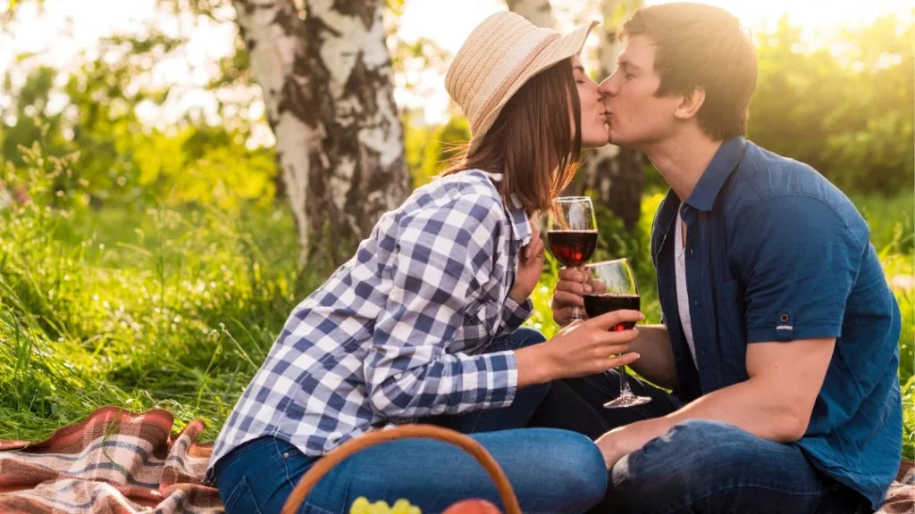 Una pareja se besa apasionadamente mientras sostiene copas de vino tinto durante un picnic romántico en un bosque o viñedo iluminado por el sol.